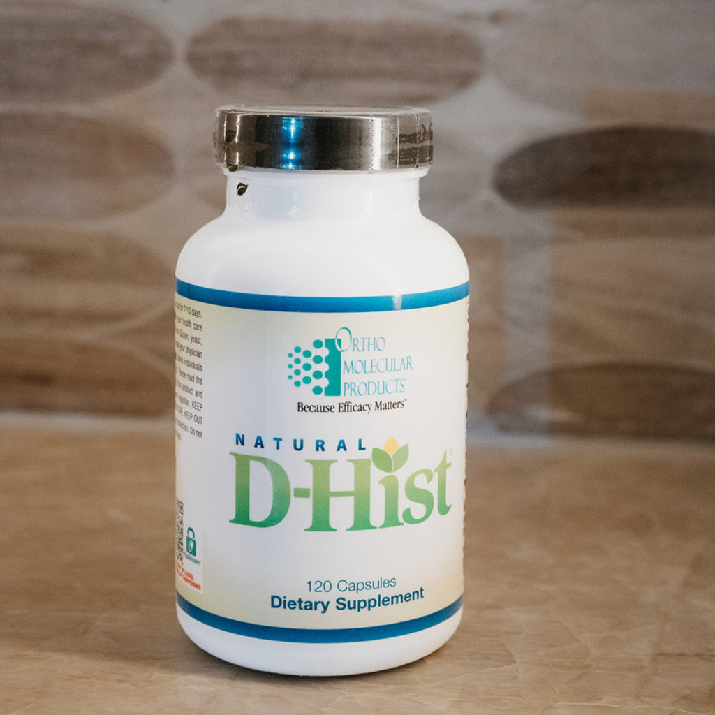 A white bottle of Natural D-Hist dietary supplement by Ortho Molecular Products, featuring a silver cap and a white label with green and blue text, set against a blurred wooden background.