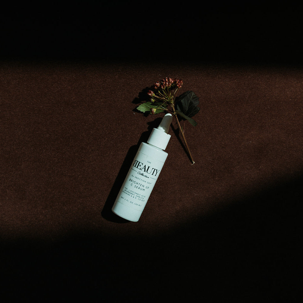 A single skincare bottle with a white pump sits on a dark brown textured surface under dramatic shadow, styled with a small dried flower sprig. The bottle label reads BEAUTY.