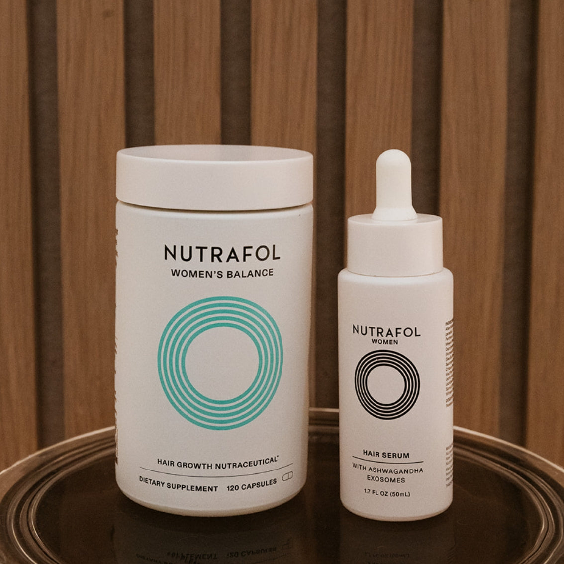 Nutrafol Women's Balance hair growth nutraceutical supplement container next to a Nutrafol Hair Serum dropper bottle, both in white with teal and black graphics, set on a dark reflective tray against a wooden slatted background.