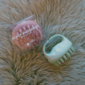 A pink and white silicone scalp massager shown next to its clear plastic packaging box on a beige faux fur background.