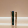A silver cosmetic eye serum bottle standing next to its dark teal and silver retail box on a reflective surface, set against a warm beige background with a horizontal light band.