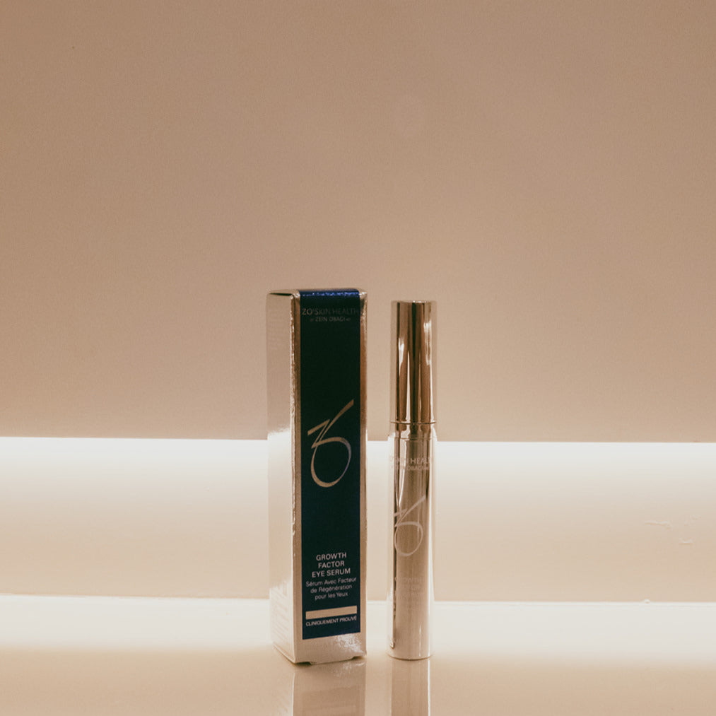 A silver cosmetic eye serum bottle standing next to its dark teal and silver retail box on a reflective surface, set against a warm beige background with a horizontal light band.