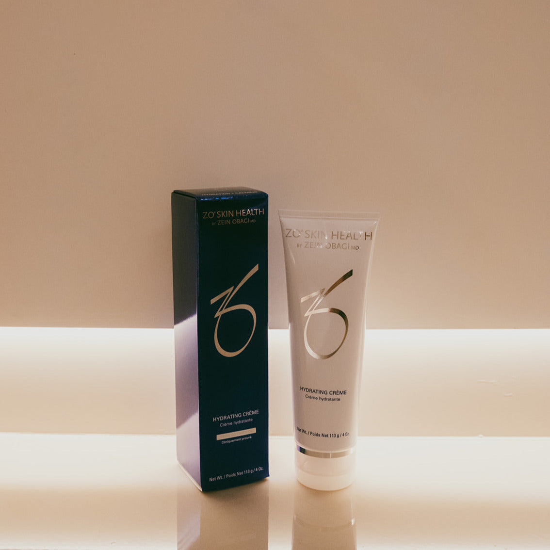 ZO Skin Health Hydrating Creme shown in a white squeeze tube next to its dark teal-blue packaging box, set against a warm beige background with a glowing horizontal light strip.