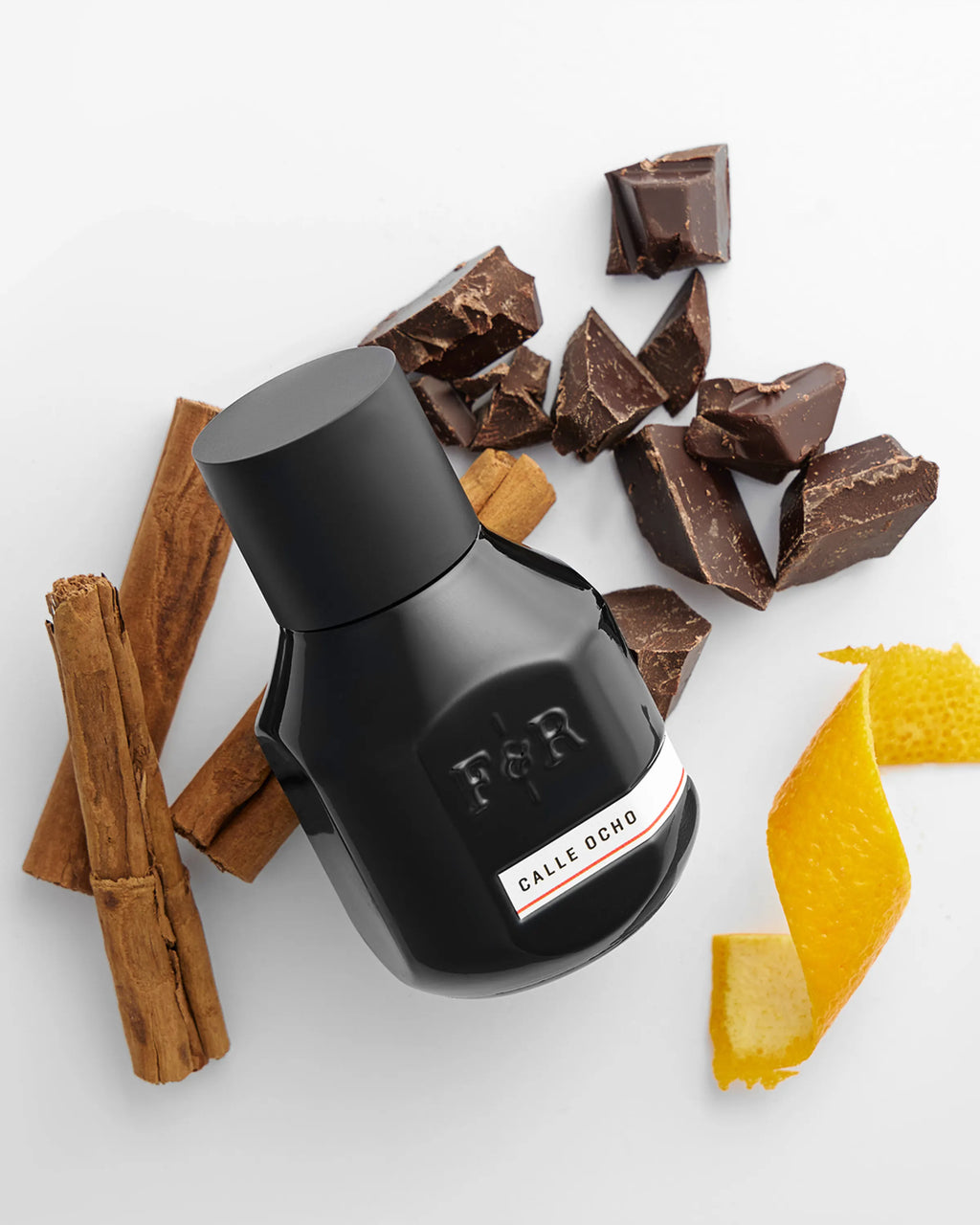 Black bottle with a label surrounded by chocolate pieces, cinnamon sticks, and orange peel on a white background